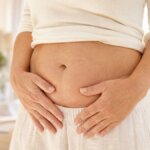Nahaufnahme von Taille und Unterbauch einer Frau mittleren Alters mit sanft aufliegender Hand in warmem Tageslicht.
