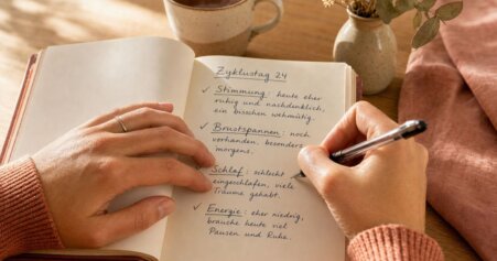 Frau in rosafarbenem Pullover schreibt in ein Zyklustagebuch mit deutschen Einträgen zu Stimmung, Brustspannen, Schlaf und Energie an Zyklustag 24 – warme Holztisch-Atmosphäre mit Teetasse und Trockenblumen.