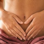 Frau hält beide Hände schützend auf den Unterbauch, rosa Tuch drapiert, warmes Seitenlicht – ruhige, körperbewusste Geste ohne Schmerzausdruck.
