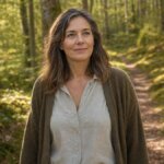 Eine Frau Mitte vierzig bis Mitte fünfzig steht auf einem Waldweg im weichen Morgenlicht, den Blick ruhig und leicht nach oben gerichtet – der Ausdruck offen und gefasst.