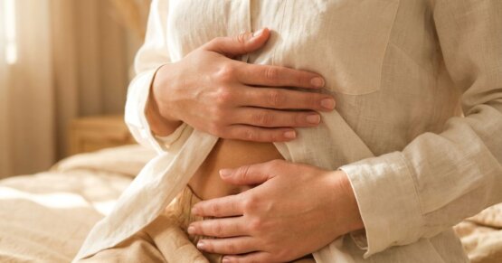 Nahaufnahme: Zwei weibliche Hände ruhen sanft auf dem Bauch, weiches Morgenlicht, cremefarbenes Leinenshirt.